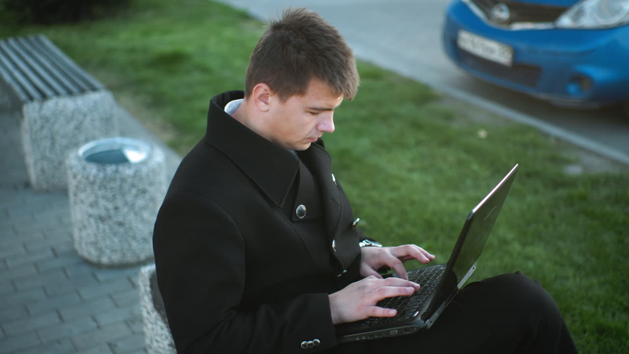 실버 버튼으로 검은색 슈트를 입은 사업가가 야외에서 노트북에 타이핑하는 동안 집중적으로 초점을 맞추고, 배경에는 녹색 풀, 보행길, 그리고 흐릿한 파란색 자동차가 있습니다.