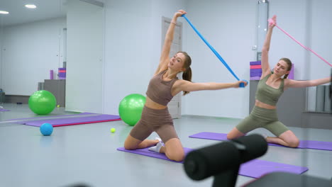 un instructor de gimnasia conduce una sesión de entrenamiento, demostrando un estiramiento de banda de resistencia mientras un aprendiz sigue sus movimientos de izquierda a derecha, con bolas de ejercicio coloridas en el fondo