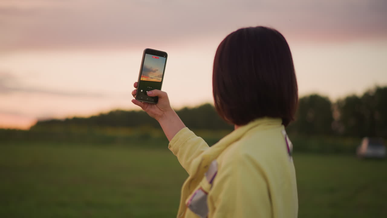 Una mujer levanta su teléfono para fotografiar una puesta de sol sobre la pradera; un smartphone encuadrando el horizonte brillante; una fotógrafa amateur y bloguera documentando una escena crepuscular; brisa suave, contraluz tenue, ambiente bucólico y cálido.