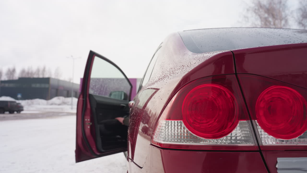 Rear view of parked red car with taillights off and water droplets on surface as driver exits and shuts door during snowy winter day with blurred buildings and distant vehicles