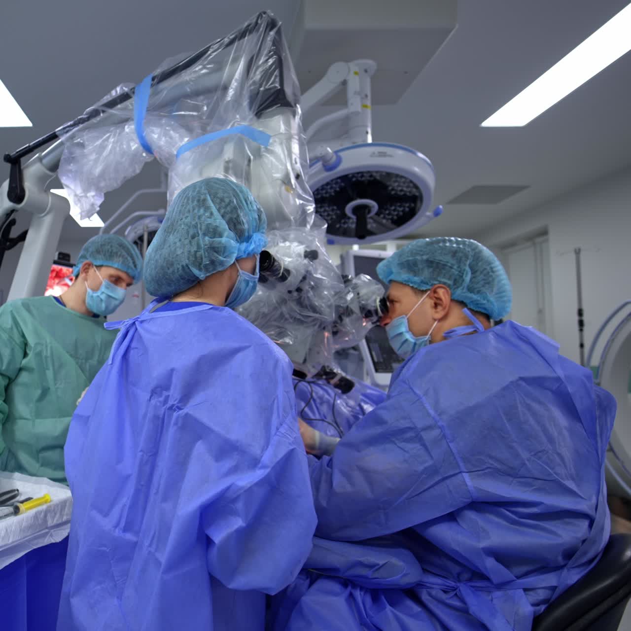 Modern neurosurgical operation conducted with the help of advanced equipment and devices. Chief surgeon and nurse look at microscope and assisting medic stands at the table with instruments