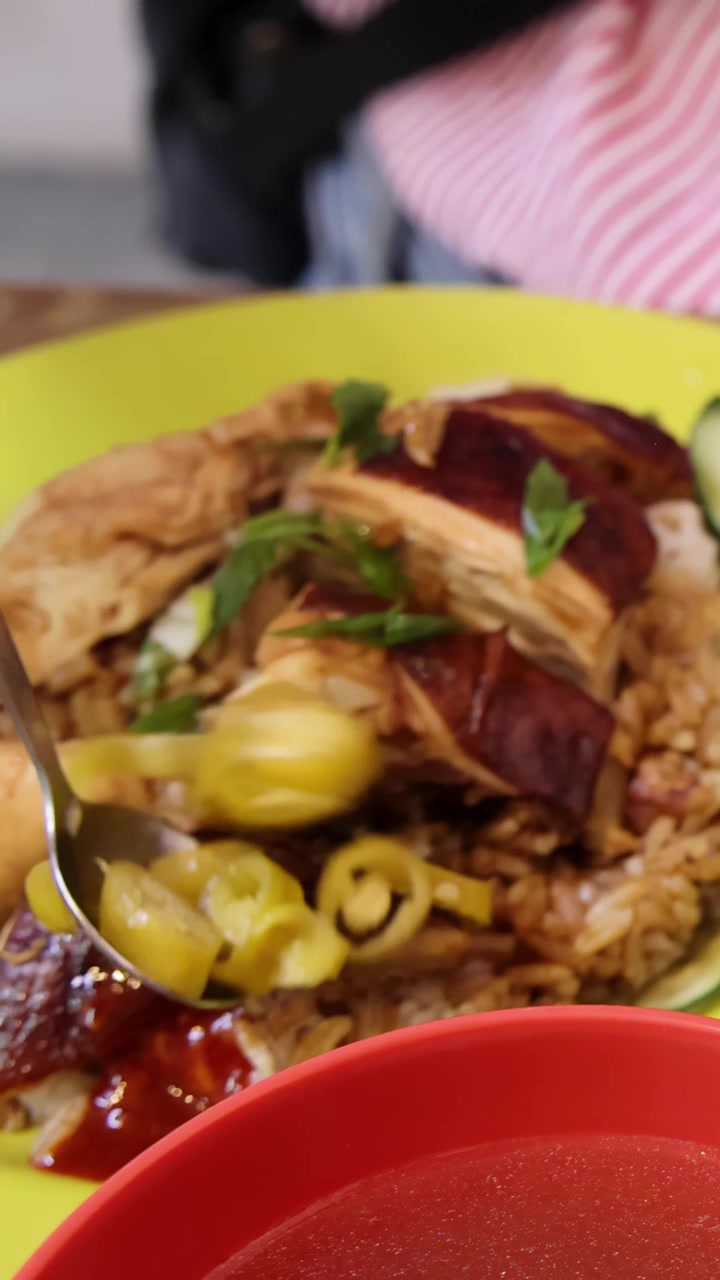 Una mano sirve rodajas de chile encurtido de un recipiente metálico sobre un plato de arroz con pollo asado y pepino, bajo una iluminación interior brillante y ángulos de cámara en primer plano