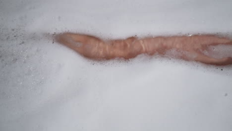 woman leg hides under soapy foam in bathtub closeup. lady washes body taking antistress bath with cosmetic products at home. woman enjoys body care routine