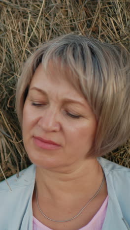 una mujer de mediana edad descansa pensativa en una bala de heno con vastas tierras de cultivo en el fondo, mira hacia la distancia, rodeada de campos ondulados y un entorno rural pacífico.