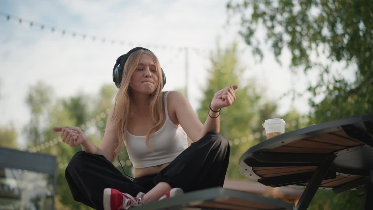 Donna caucasica con cuffie seduta all'aperto, che si muove a ritmo di musica su un tavolo da picnic, movimenti giocosi delle mani, scarpe da ginnastica rosse in vista, tazza di caffè sul tavolo, lucine sospese, luce solare calda filtrata