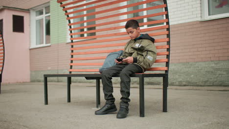 young student seated on modern bench playing game with phone during school breaktime bag resting by side flag visible through window waving gently in wind