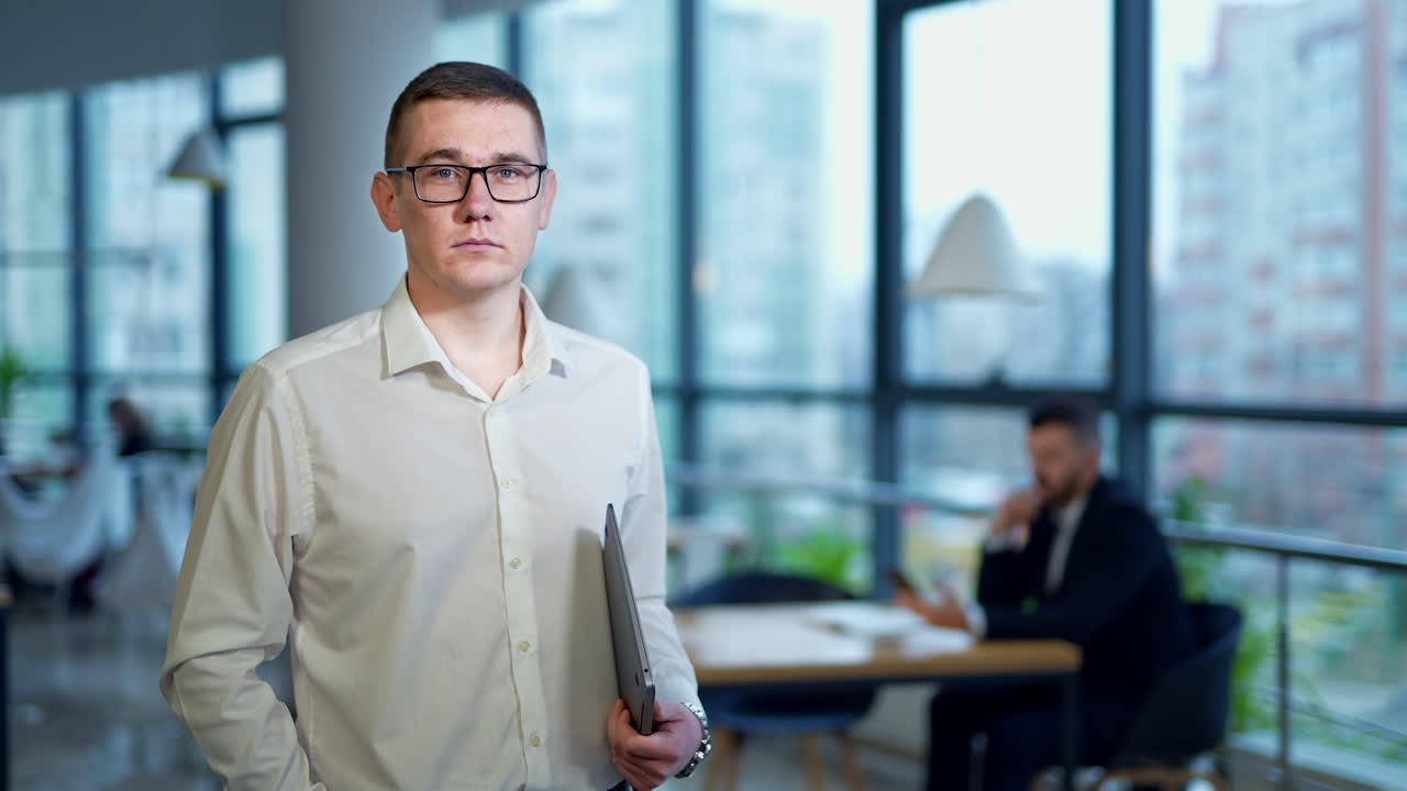 Caucasian man wearing glasses and holding a laptop in the office. Male employee portrait standing against the panoramic windows backdrop.