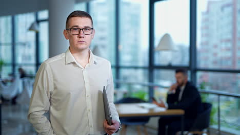 caucasian man wearing glasses and holding a laptop in the office. male employee portrait standing against the panoramic windows backdrop.