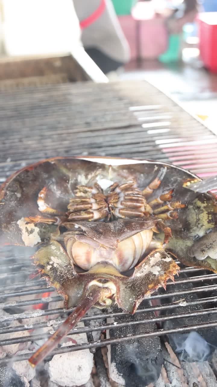 A horseshoe crab cooks on a metal grate over smoking charcoal in a vertical shot