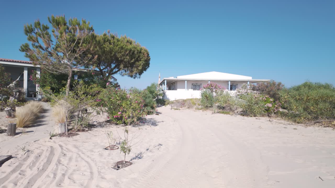 Whitewashed vacation homes surrounded by lush vegetation bask in the sun on the sandy shores of Ilha da Armona, a serene island retreat near Olhao, Portugal