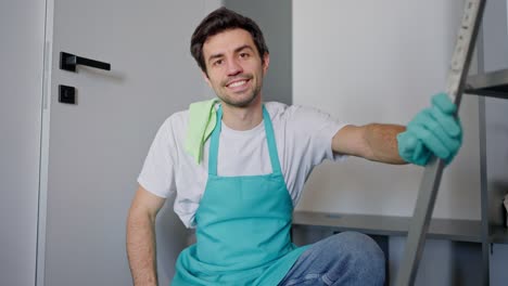 retrato de un confiado y feliz limpiador masculino en un delantal azul arrojando un trapo sobre su hombro y posando mientras está sentado en el suelo entre herramientas de limpieza y una escalera en un apartamento moderno