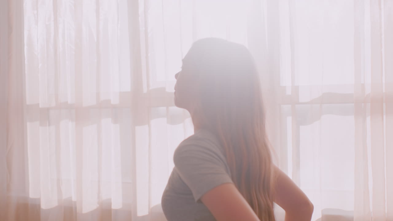 Young woman standing beside window stretches shoulders backward with eyes closed, soft light filling room through sheer curtains, serene expression suggesting peaceful morning routine