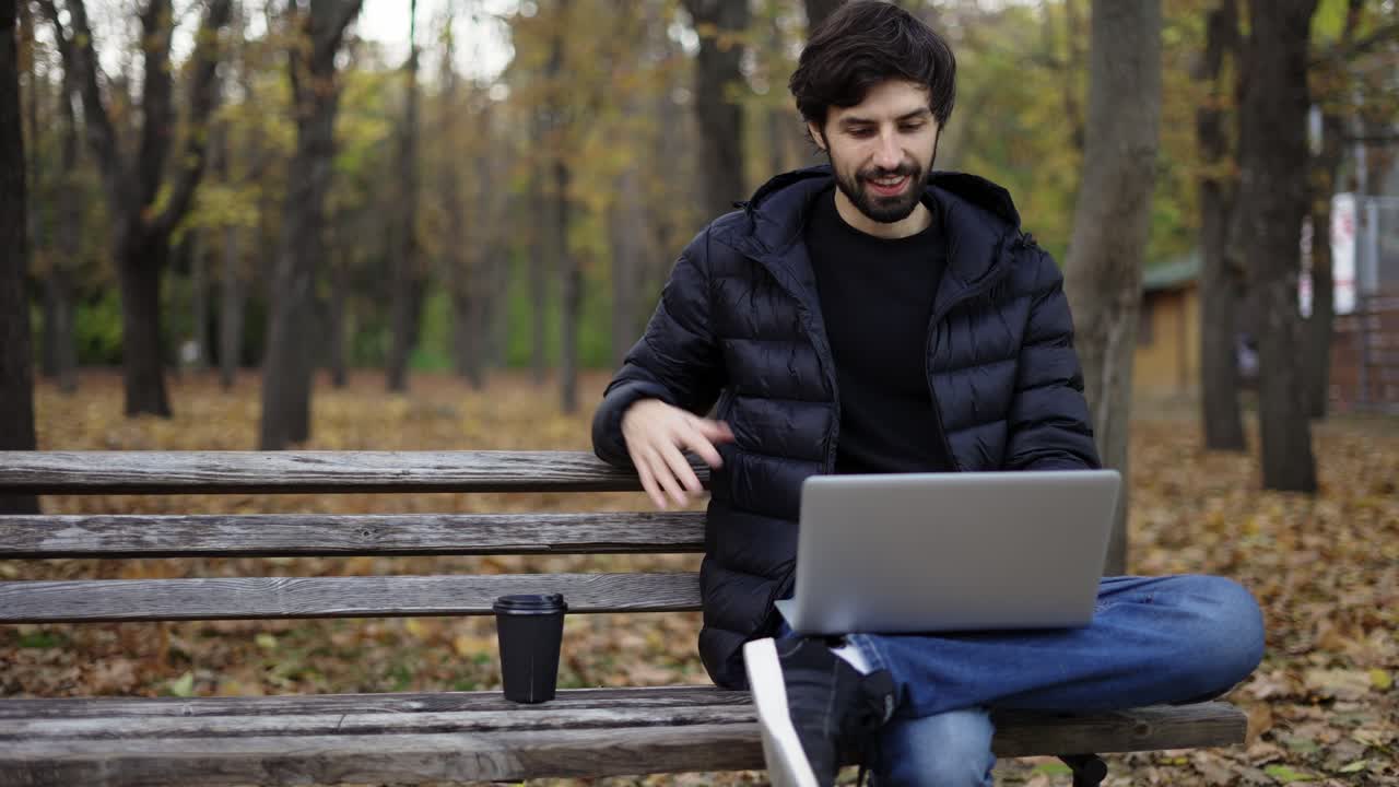 hombre sonriente sentado casualmente en un banco en un parque público, usando una computadora portátil para una llamada de video