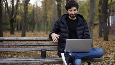 smiling man sitting casually on bench in public park, using laptop for video call