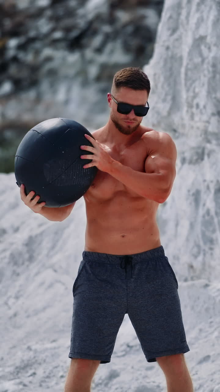 Bodybuilder training with a heavy ball in white mountains. Strong guy wearing black sunglasses doing exercises with a ball over his head outdoors in summer. Vertical video