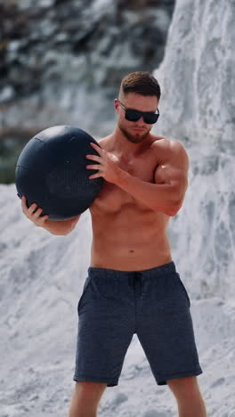 bodybuilder training with a heavy ball in white mountains. strong guy wearing black sunglasses doing exercises with a ball over his head outdoors in summer. vertical video
