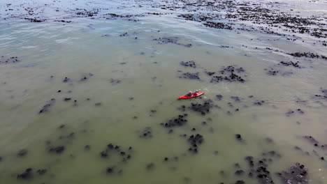 pescando en una canoa roja brillante que contrasta con el agua sucia del océano llena de algas, un dron aéreo que sigue al remero