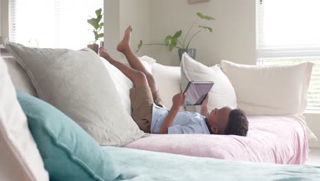 happy african american boy lying on sofa with legs up using tablet at home, slow motion