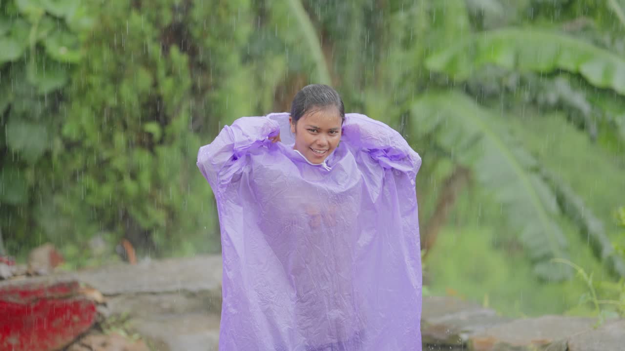 Indian rural girl laughing and looking up at the sky as rain falls, enjoying monsoon in a raincoat, 4k video