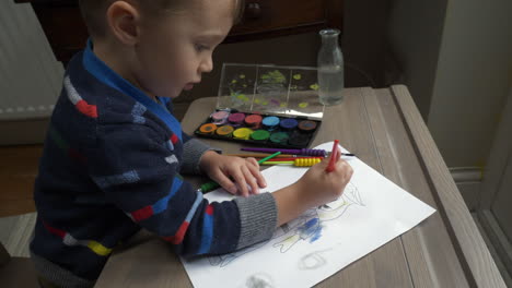 a mid shot of a small boy at a desk painting the face of a pre drawn pirate figure in black watercolour