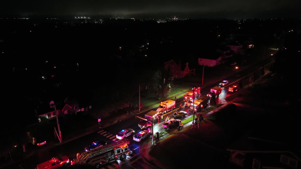 Police officer, firefighter and ambulance helping injured people of car accident at night. American neighborhood with intersection. Aerial establishing shot.