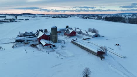 Sonnenaufgang-über-Einem-Verschneiten-Bauernhof-Mit-Roten-Scheunen,-Silo-Und-Kahlen-Bäumen