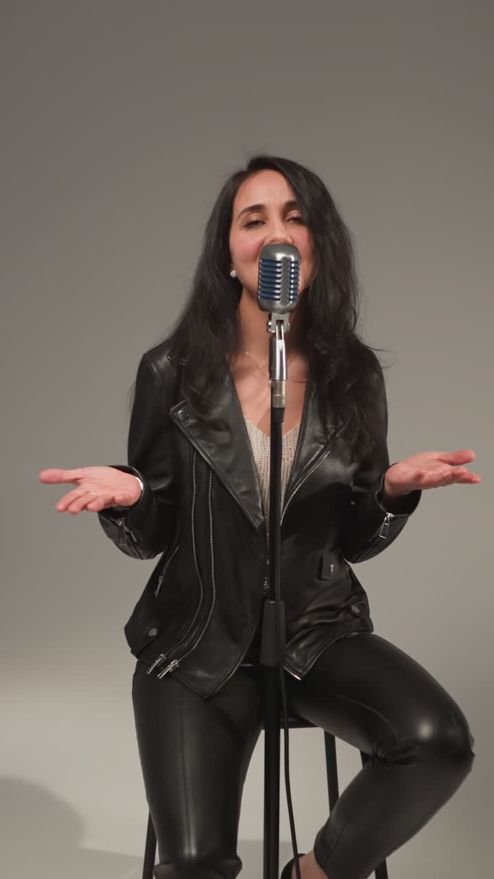 una hermosa vocalista con una chaqueta de cuero negra, sonriendo y cantando con los ojos abiertos y las manos moviéndose expresivamente, de pie en un micrófono vintage en un taburete alto, actuando contra un fondo gris