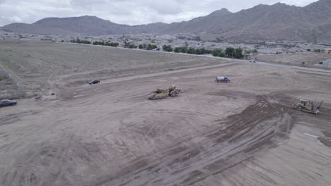 Un-Dron-Rodea-Con-Gracia-Una-Traílla-Elevadora-Estacionada-En-Medio-De-Un-Lote-En-Un-Posible-Sitio-De-Construcción-De-Una-Casa,-Ofreciendo-Una-Visión-De-Otra-Maquinaria-Pesada-Trabajando-Diligentemente