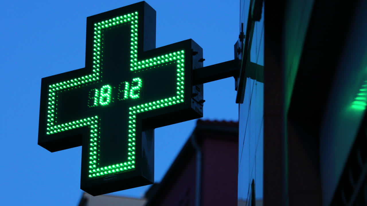 Riflesso di una farmacia e del suo simbolo internazionale luminoso (una croce verde con orologio lampeggiante al centro) appeso all'angolo di un edificio, ripreso in 4k a 60fps.