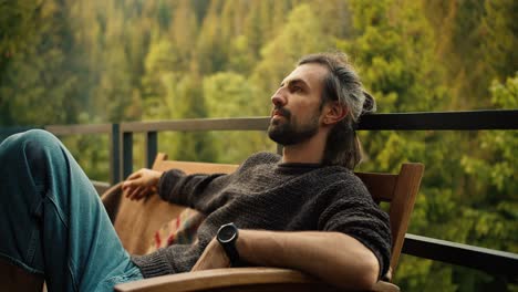 a young brunette man is resting, sitting on a sofa against the backdrop of mountains and coniferous forest