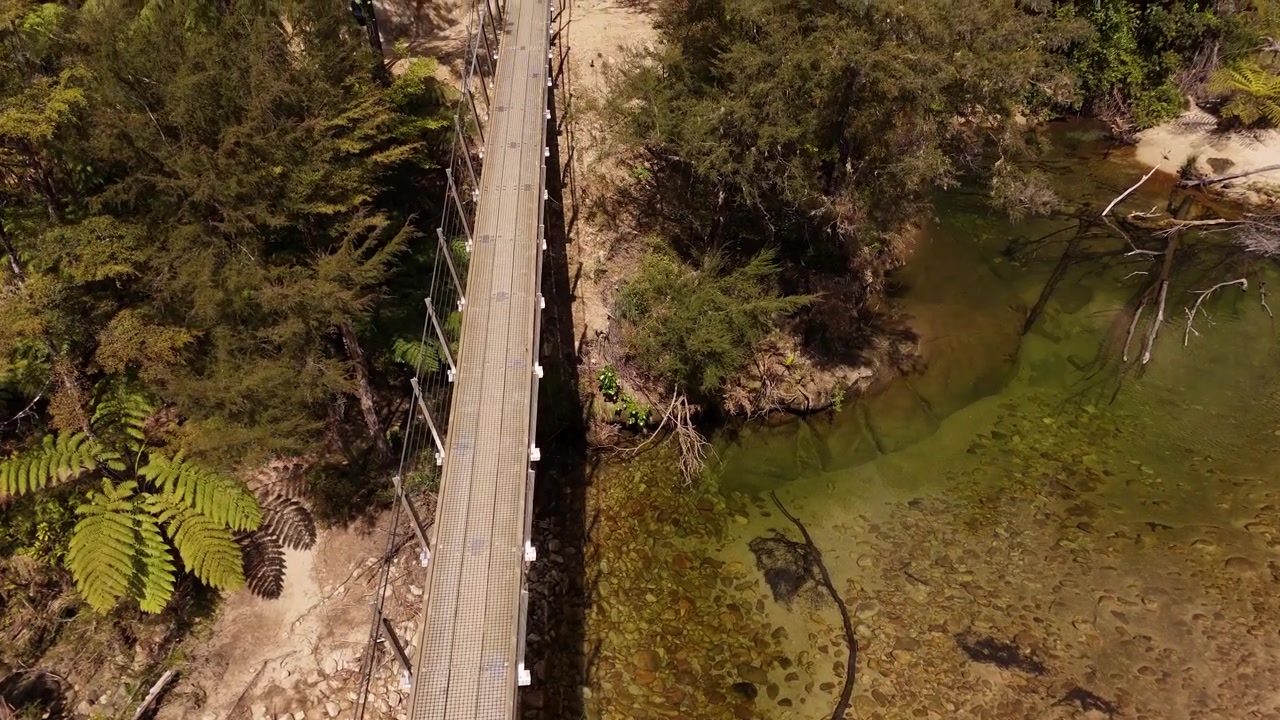 Lang hængebro for fodgængere over flod i frodig urskov i Abel Tasman Nationalpark, New Zealand. Luftfoto ovenfra, fremad, i slowmotion.