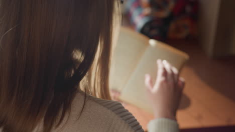 close up rear view of lady reading book by window as sunlight casts soft shadows across wooden surface, gentle light enhances calm indoor ambiance with blurred pages