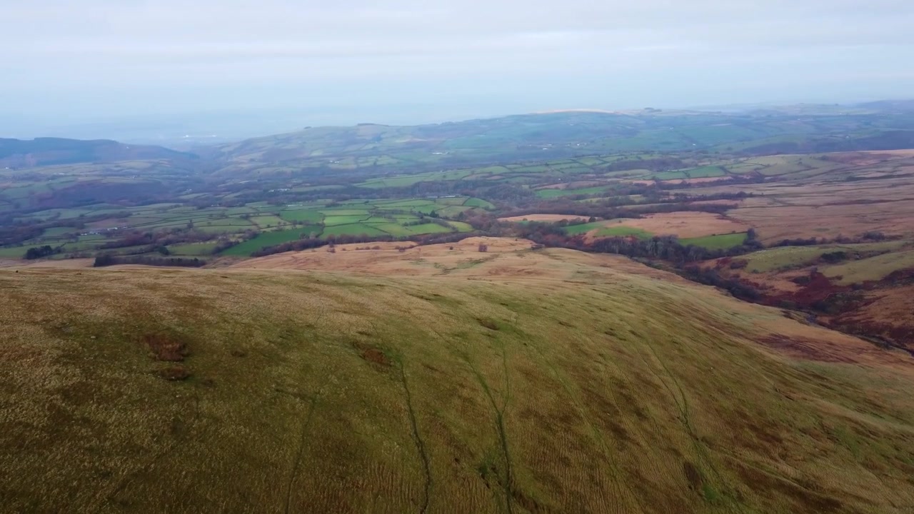 Ampia e veloce vista aerea con drone attraverso una valle gallese con paesaggio di brughiera dei Brecon Beacons e campi coltivati in lontananza velata 4K.