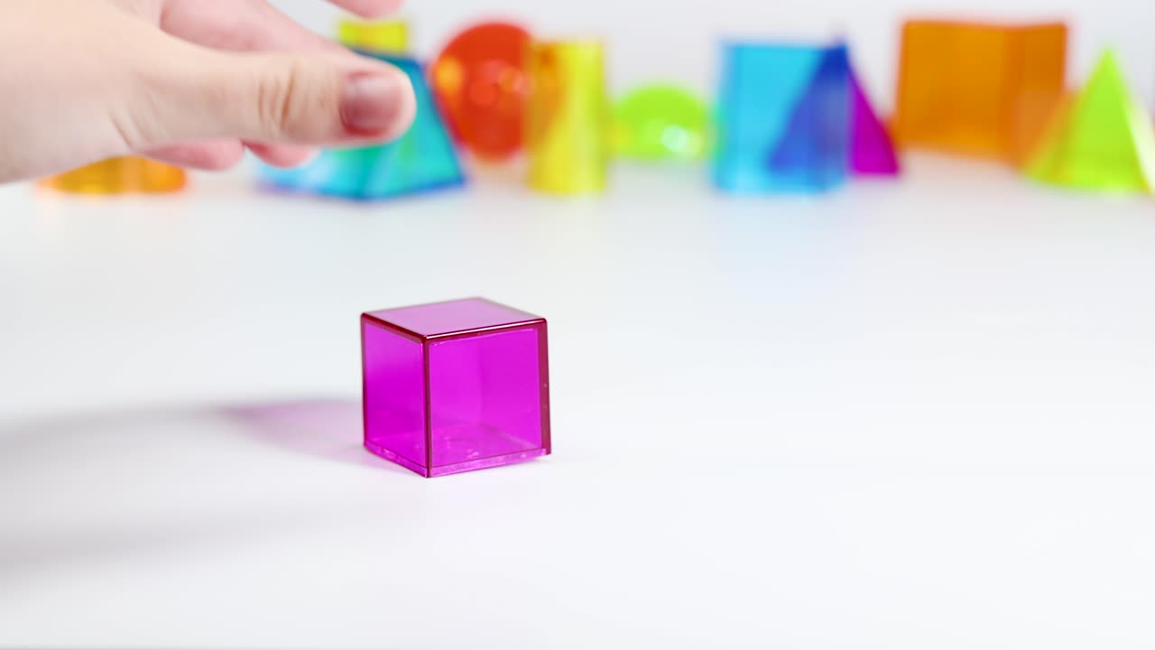 A hand interacts with a pink cube among colorful 3D geometric shapes on a white surface, under bright lighting