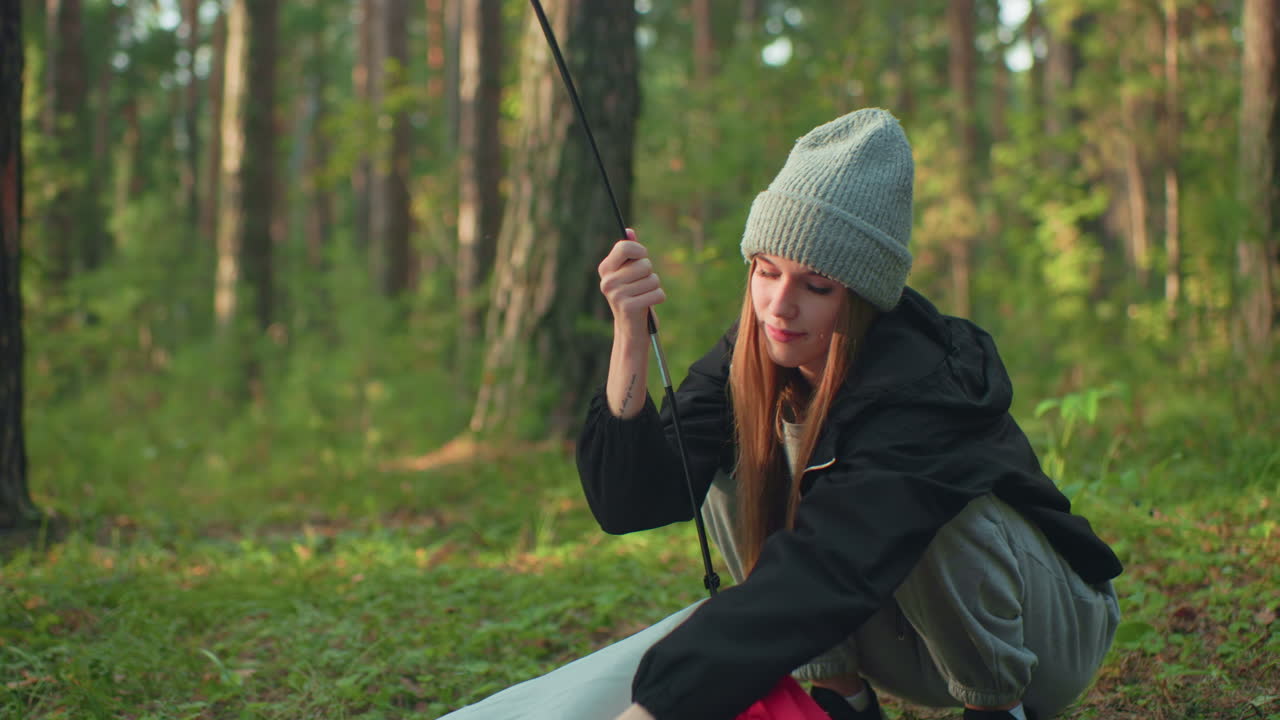 Young girl squats in forest fixing tent fabric hook onto flexible pole, maneuvering clip through ring while balancing gear and adjusting angle under warm sunlight