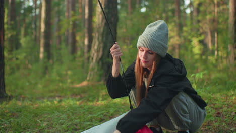young girl squats in forest fixing tent fabric hook onto flexible pole, maneuvering clip through ring while balancing gear and adjusting angle under warm sunlight