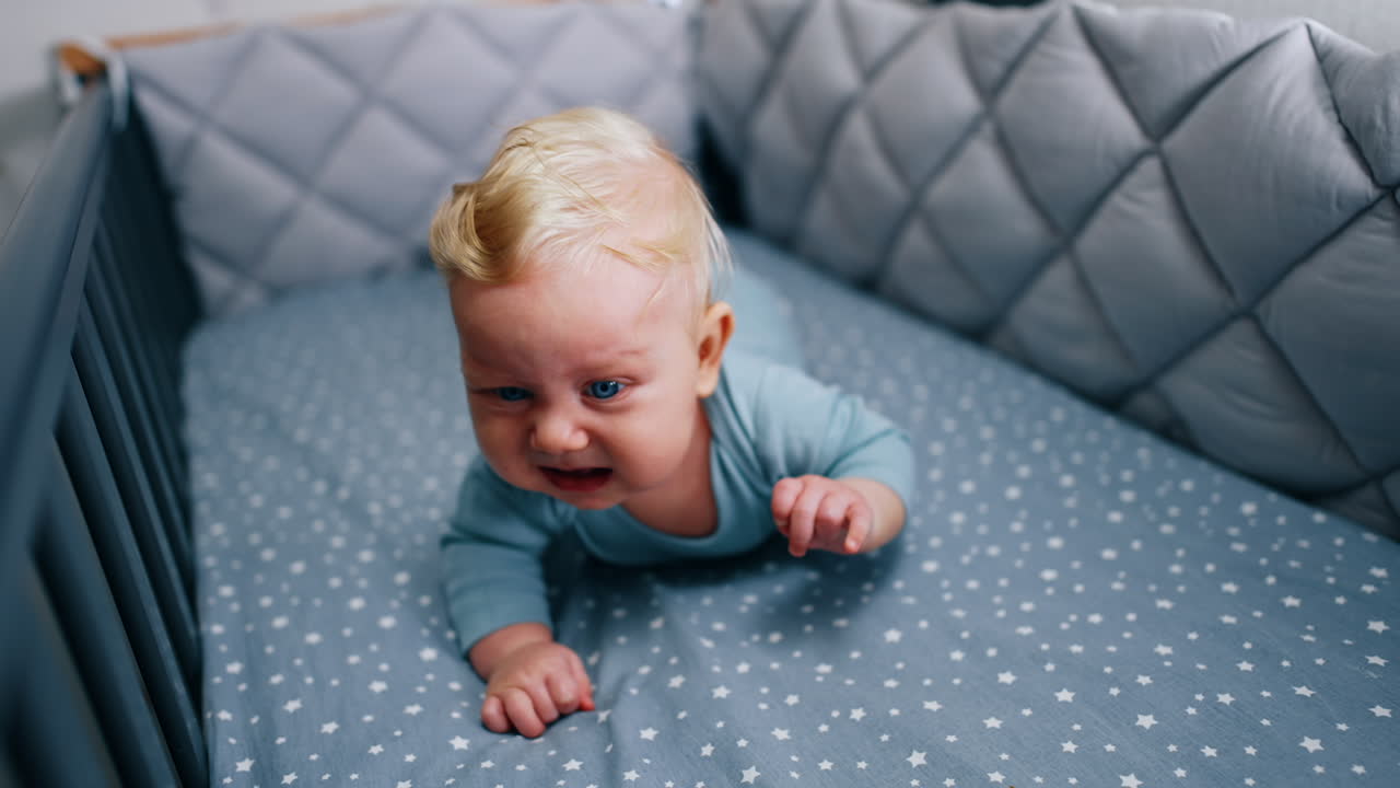 Caucasian infant baby lies in comfortable crib. Blond kid with blue eyes starts to cry. Close up.