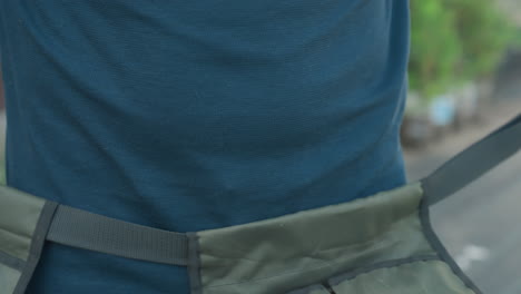 close up man retrieving apron belt from wooden crate on blue drum, lifting strap with buckle, preparing wear around waist near garden tools, gloves, trowel, fork, sprayer, house yard