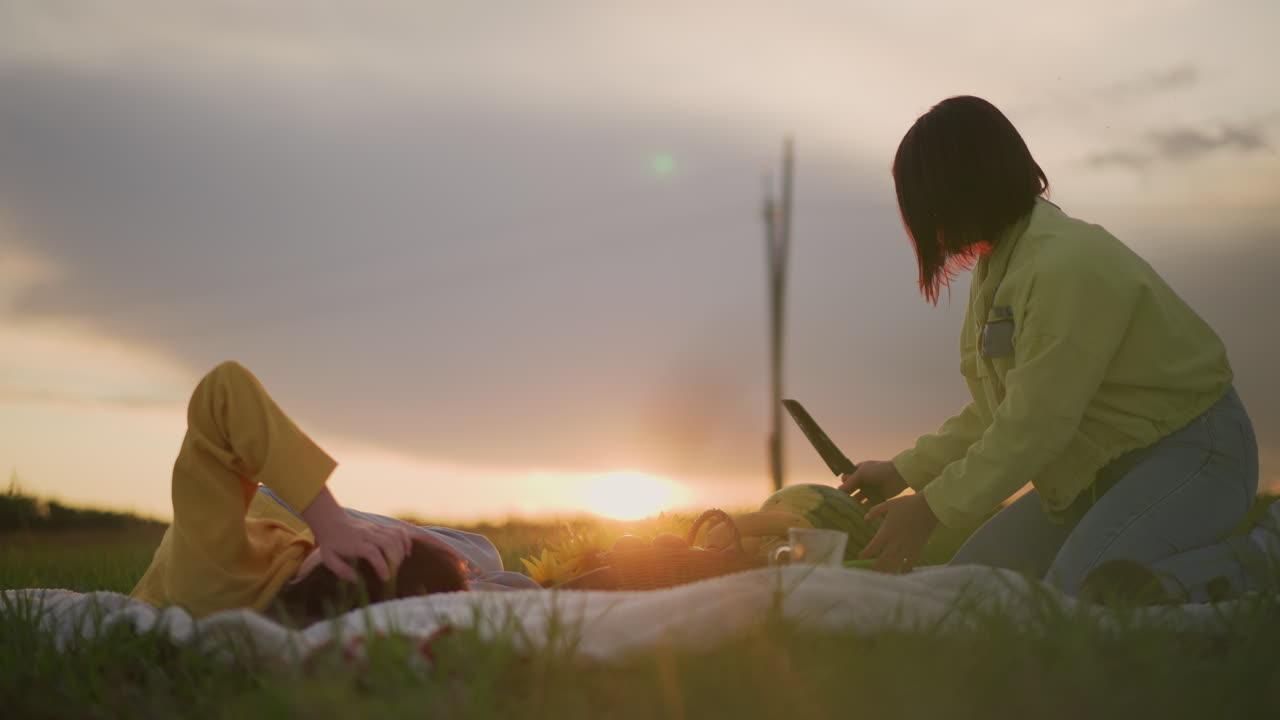 해 질 녘 피크닉 담요 위에서 친구 옆에 무릎을 꿇고 앉아 따뜻한 햇살을 받으며 즐거운 시간을 보내는 백인 여성, 장난기 넘치는 몸짓으로 간식을 권하고, 친구는 노란색 후드티를 입고 편안하게 누워 있는 자연스러운 모습
