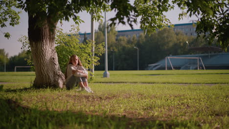 frau sitzt auf grasbewachsenem boden unter einem baum mit den armen über die beine gefaltet, tief im gedanken, das sonnenlicht reflektiert von ihr, mit hintergrund einschließlich stangen und fernem stadion