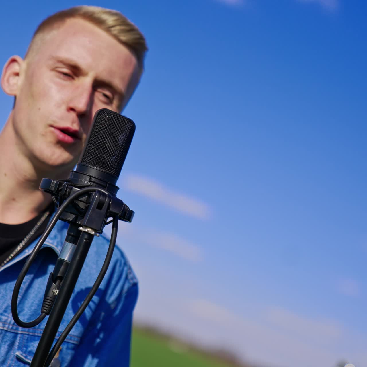 Young blond male in jeans jacket comes up to a microphone starting to sing. Rock band performing in the farmland on sunny day
