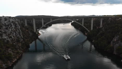Drone-areal-lift-shot-of-sailing-boat-driving-under-car-bridge-in-fjord-in-Croatia