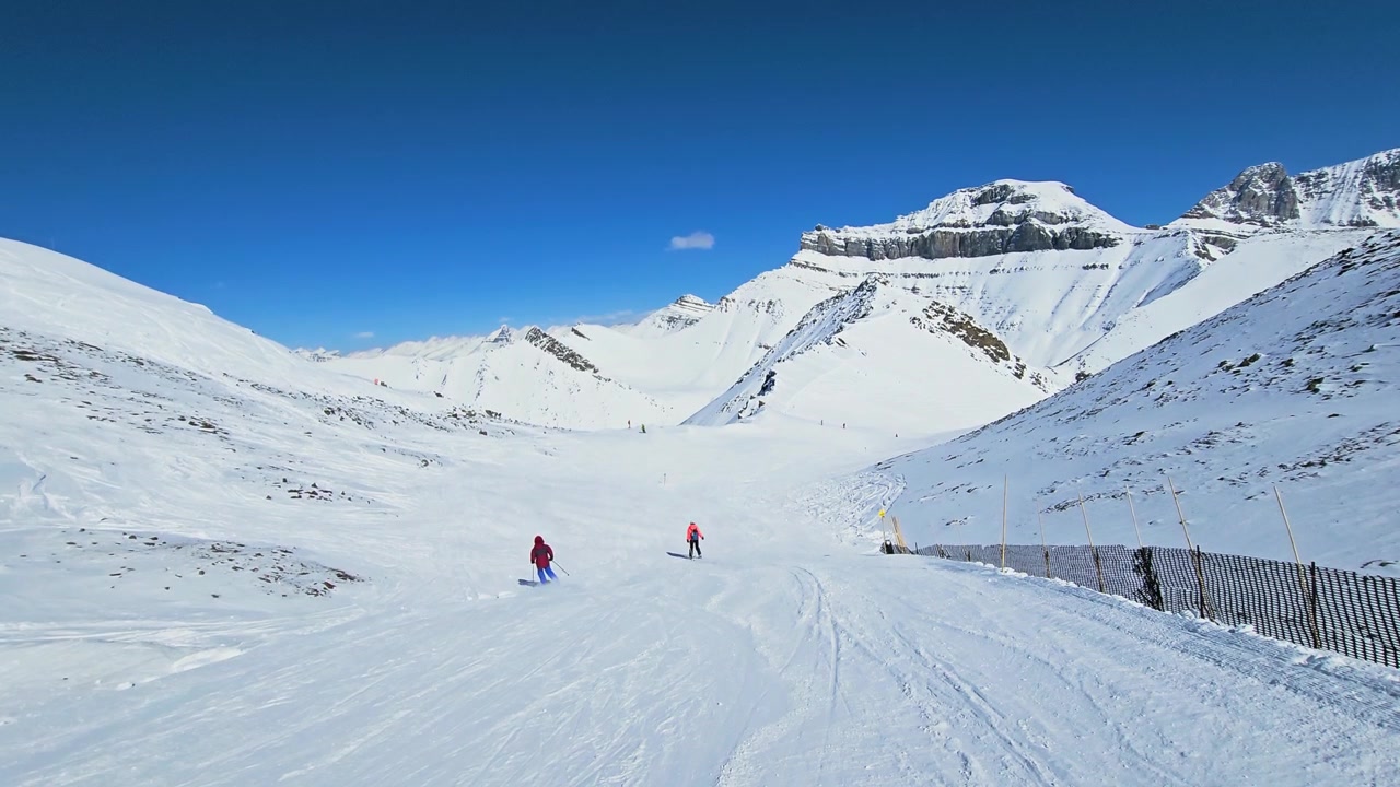 Skiers glide downhill at Aspen Snowmass, surrounded by white slopes, distant Colorado peaks, and a clean blue sky.