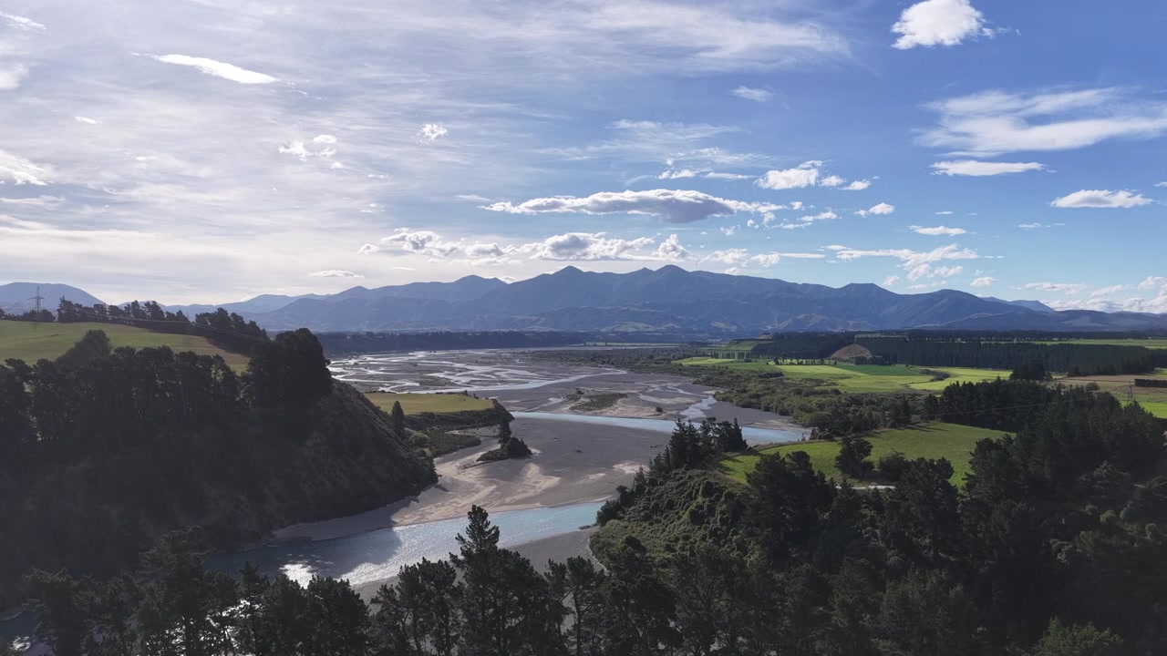 Vista aerea di canali fluviali intrecciati che serpeggiano attraverso paesaggi agricoli e forestali sull'Isola del Sud della Nuova Zelanda, sotto cieli vasti e catene montuose distanti.