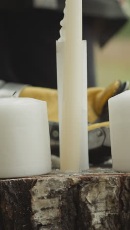 White candles stand on dry stump on lawn. Couple of people in medieval clothes stand looking at objects. Female hand puts knight gauntlet closeup on blurred background