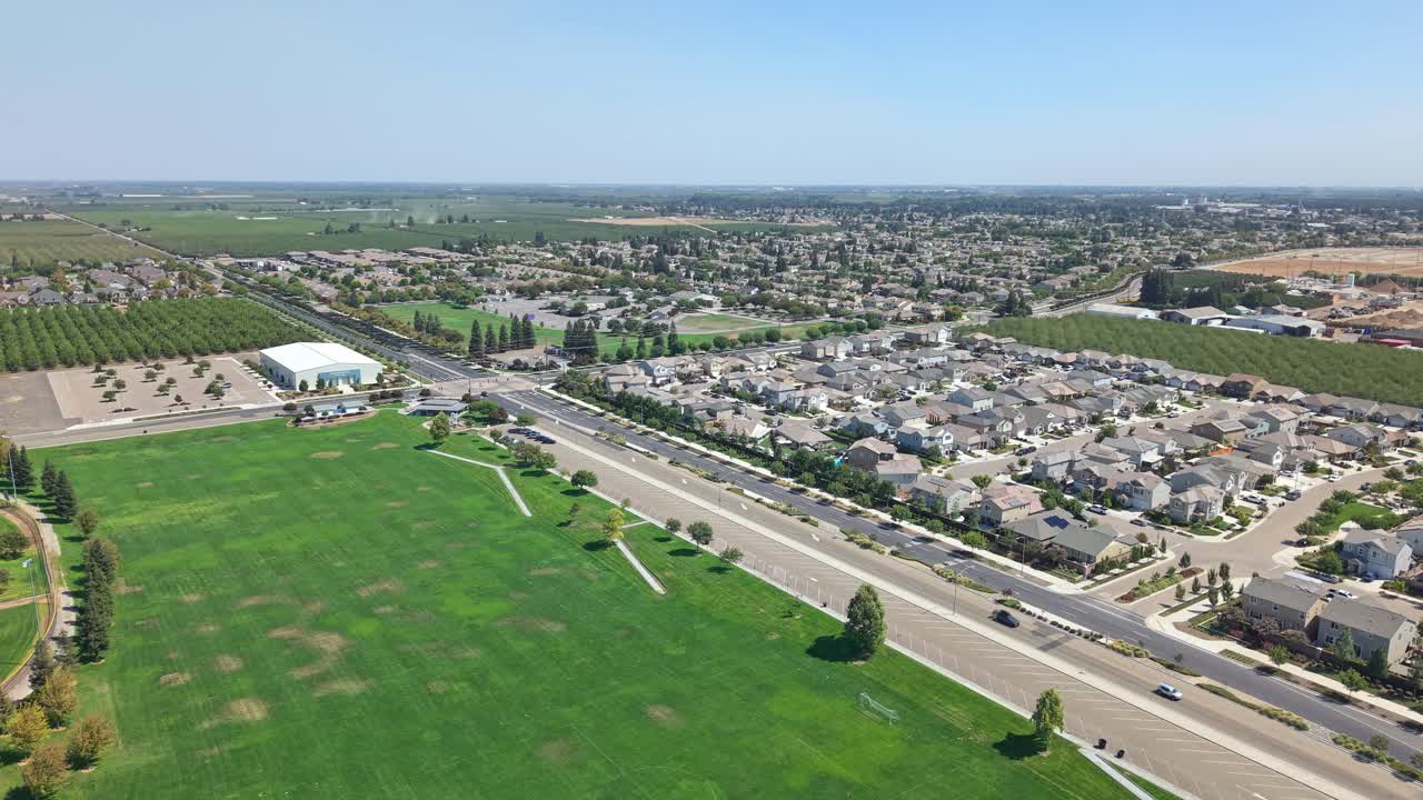 High drone shot rotating above grassy park and road dividing Ripon residential area, showing houses, traffic, farmland, and large recreational building