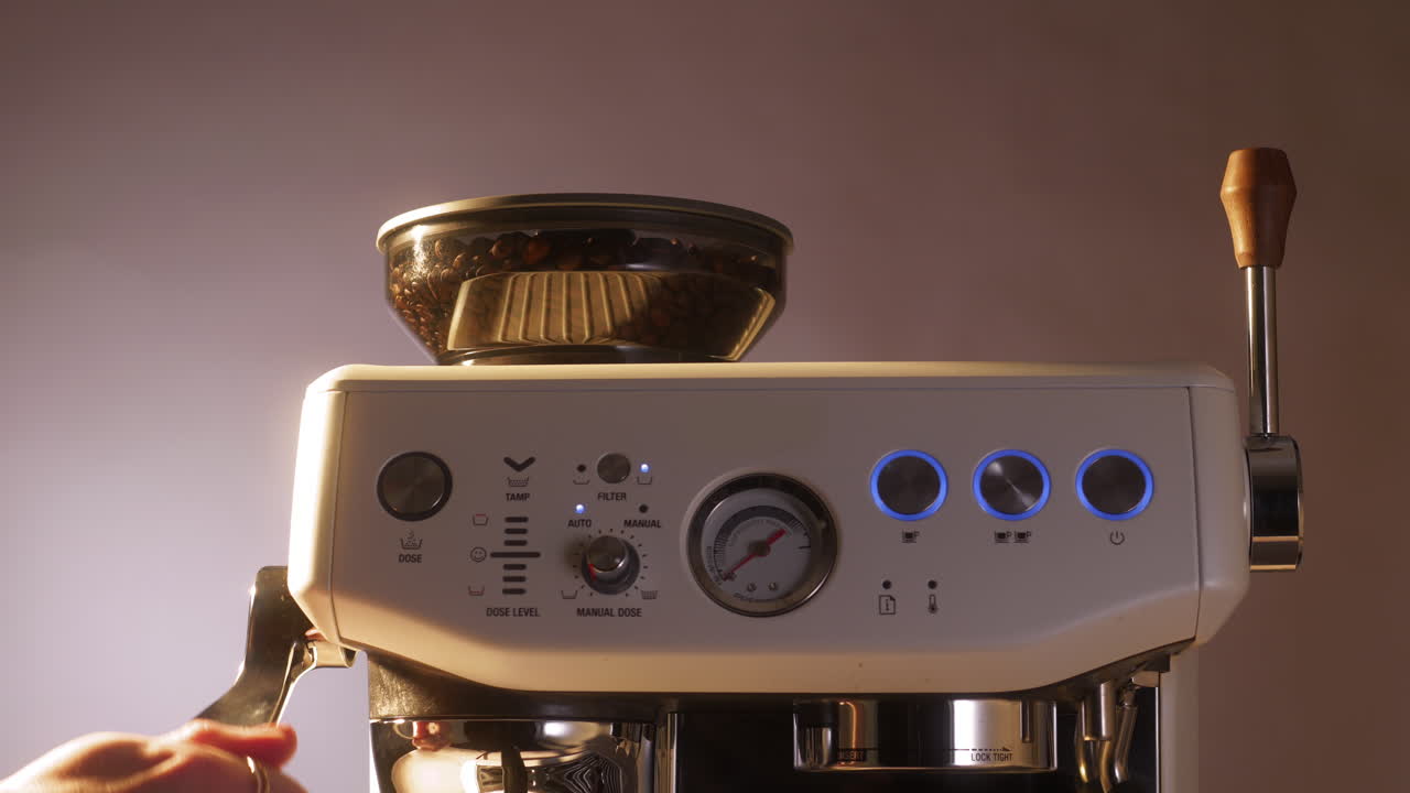 Primer plano de una mano presionando el portafiltro contra la palanca del molinillo en una elegante máquina de espresso, con granos visibles en la parte superior y botones iluminados que indican el modo de preparación de café activo.