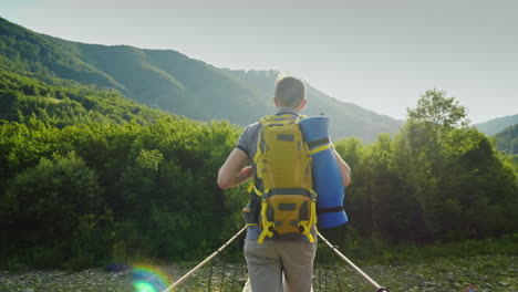 a traveler with a backpack walks along a wobbly bridge over a mountain river back view 4k video