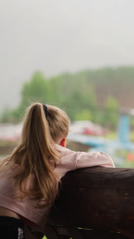 bored little child leans on large old log railing and looks at helicopter with rotating propeller on terrace of rural cafe on rainy day slow motion