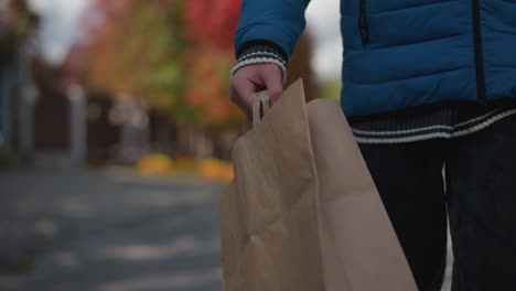 partial view of someone walking in a residential area carrying a paper bag, framed by autumn trees, houses, and a parked car in the soft-focus background